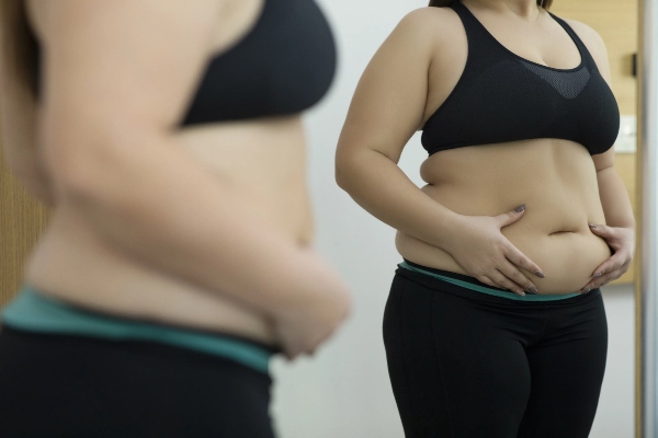 clínicas de abdominoplastia en Bilbao - mujer obesa con ropa deportiva mirándose en el espejo mientras coloca su manos sobre la panza