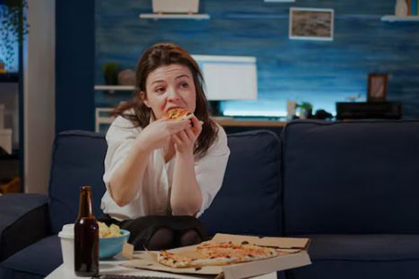 Joven con estilo de vida sedentario y con malos hábitos alimenticios come un trozo de pizza frente al televisor